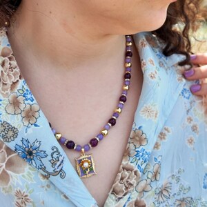 Daylight close-up against skin showing lilac and magenta-purple round beads spaced with gold-tone balls, the enamel pendant and its pearl-white flower centre hanging in the shirt opening.