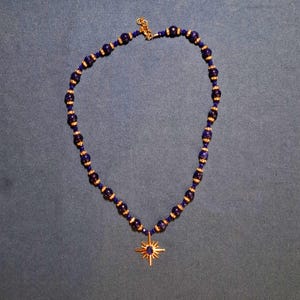 Deep navy-blue beaded necklace laid in an oval on grey-blue fabric, round stone beads alternating with gold-tone crystal rondelles and small faceted beads, a gold starburst pendant hanging at the bottom.