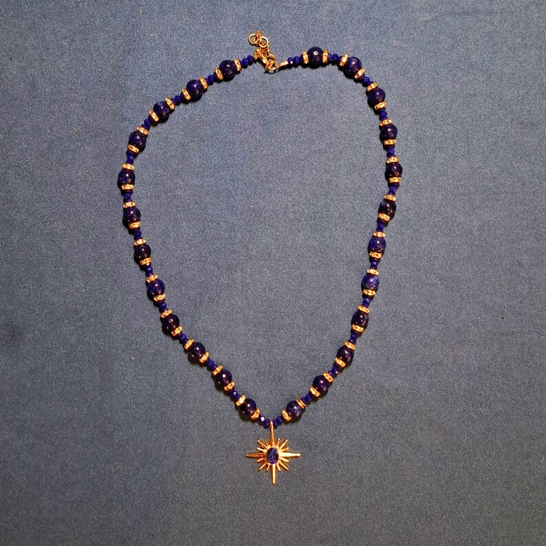 Deep navy-blue beaded necklace laid in an oval on grey-blue fabric, round stone beads alternating with gold-tone crystal rondelles and small faceted beads, a gold starburst pendant hanging at the bottom.