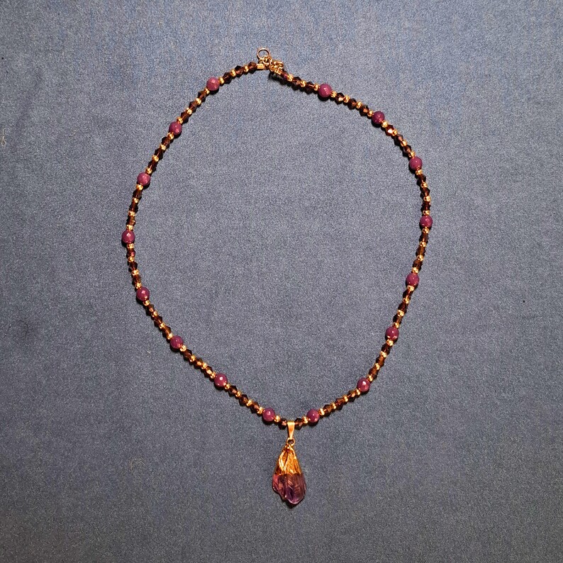 Beaded necklace laid in a circle on grey-blue fabric: round plum-purple beads alternating with faceted bronze bicones and tiny gold-tone spacers, a raw violet crystal point in a gold cap hanging at the front.