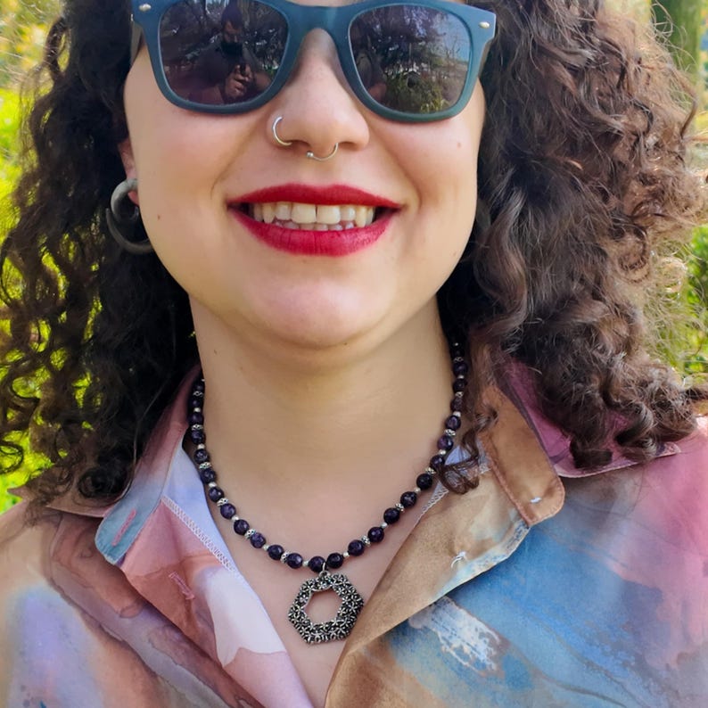 The beaded strand worn at the base of the throat over a pastel printed shirt, deep violet beads catching daylight and the flower-filigree hexagon pendant resting flat on the chest.