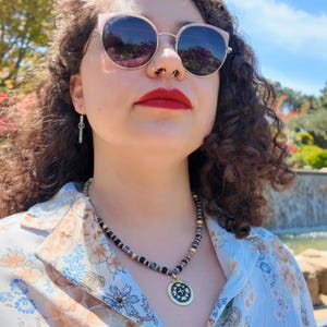 Close crop of the necklace on the chest: matte navy beads, a few blue-and-white banded ones and pale grey beads, gold and rhinestone spacers between them, and the gold medallion with its floral relief on a dark ground.