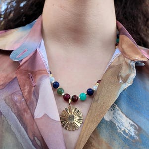 Daylight close-up of the front of the necklace: a green bead with darker mottling, a navy bead, a bright teal bead and two wine-red beads framing the bail of the gold sunburst pendant.