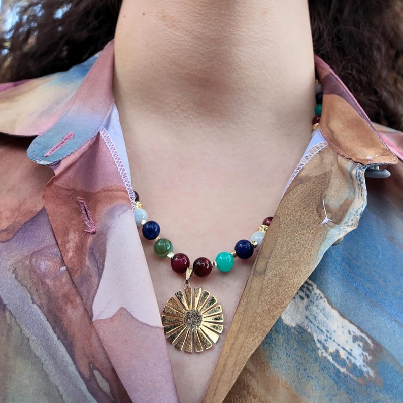 Daylight close-up of the front of the necklace: a green bead with darker mottling, a navy bead, a bright teal bead and two wine-red beads framing the bail of the gold sunburst pendant.