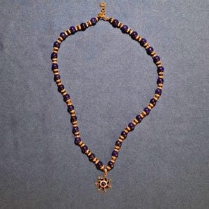 Deep navy-blue round stone beads laid out in an open loop on grey-blue cloth, a small gold rondelle sparkling in every gap, with a gold sun pendant and its blue centre stone resting at the bottom of the strand.