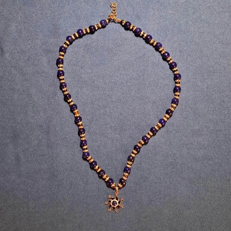 Deep navy-blue round stone beads laid out in an open loop on grey-blue cloth, a small gold rondelle sparkling in every gap, with a gold sun pendant and its blue centre stone resting at the bottom of the strand.
