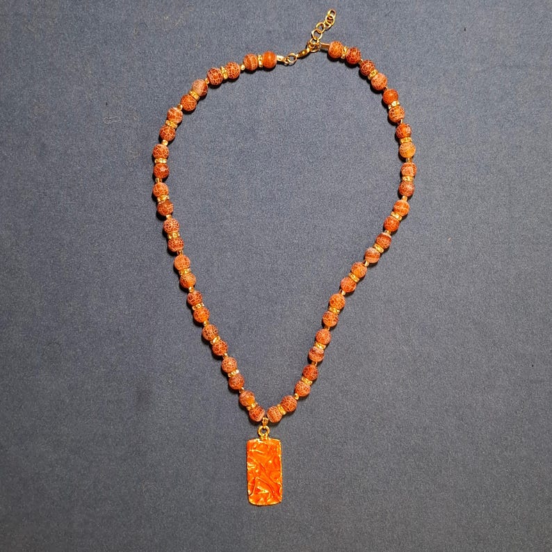 Flat-lay on grey-blue cloth: a single strand of rust-orange crackle beads with gold-tone and rhinestone rondelles, a lobster clasp and extender chain at top, and a rectangular coral pendant hanging at the front.