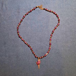 Deep plum faceted stone beads laid in an oval on grey-blue fabric, separated by tiny gold-tone spacers and dark bronze bicones, with a claw-set purple nugget and gold spike pendant at the bottom.