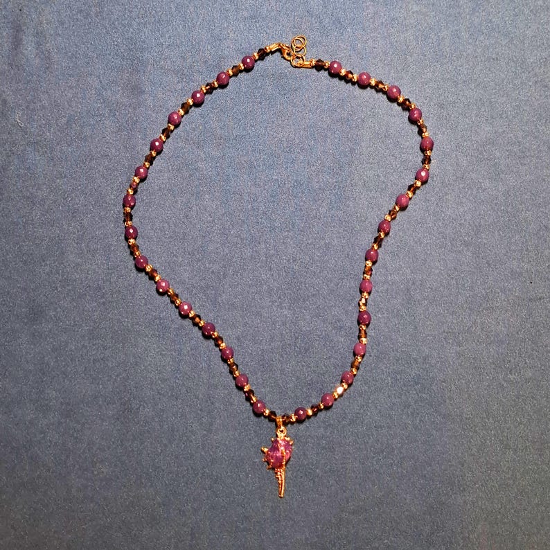 Deep plum faceted stone beads laid in an oval on grey-blue fabric, separated by tiny gold-tone spacers and dark bronze bicones, with a claw-set purple nugget and gold spike pendant at the bottom.