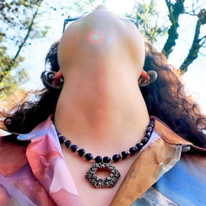Close view in strong sun showing the faceted purple beads and the little round and daisy spacers between them, with the flower-patterned hexagon pendant hanging just below the collarbones.