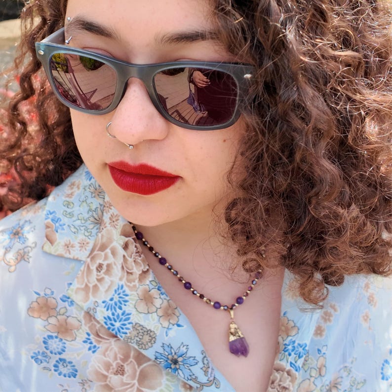Daylight close-up of the necklace against a floral blouse, showing the alternating round purple beads and faceted bronze bicones and the raw violet crystal drop below.