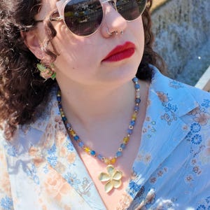 The strand worn in bright daylight, alternating denim-blue and ochre beads catching the sun, with the petal pendant hanging just below the collar of a soft blue printed blouse.