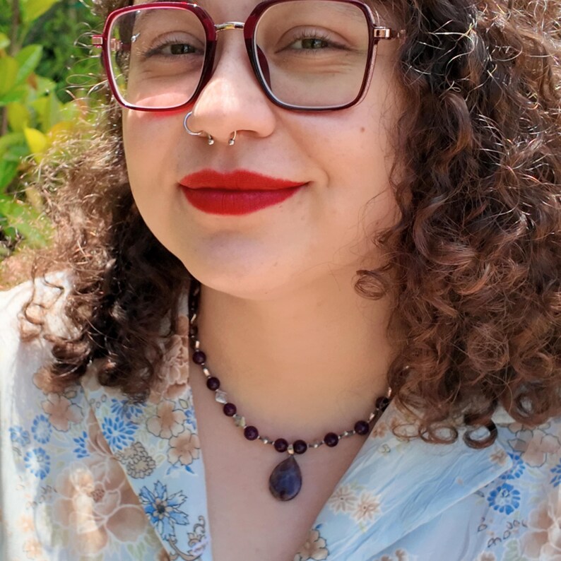 Close daylight portrait with the necklace sitting at the base of the neck — round deep purple beads, bright silver-tone spacers and a smooth teardrop pendant resting against a pale blue floral shirt.