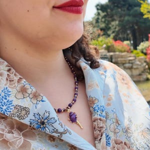 Side-on daylight shot showing the strand curving under the collar of a floral blouse, the purple beads catching sun and the spiked pendant hanging free at the front.