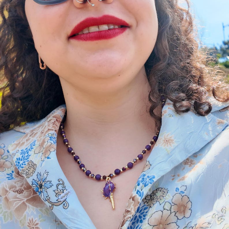 Close view of the strand in daylight: deep violet faceted rounds with gold-tone spacers and smoky bicones, ending in a rough purple stone clasped by gold prongs above a slim gold spike.