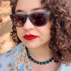 Head tilted back beside a tree trunk, the strand follows the base of the throat; in sunlight the faceted beads read forest green, sea-blue and olive, each about the width of a shirt button.