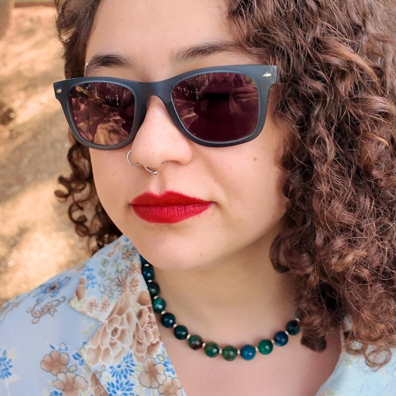 Head tilted back beside a tree trunk, the strand follows the base of the throat; in sunlight the faceted beads read forest green, sea-blue and olive, each about the width of a shirt button.