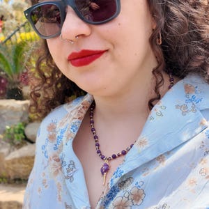 The necklace worn against a pale blue floral shirt by water, the row of small purple beads sitting close to the neck with the gold-tipped purple pendant below.