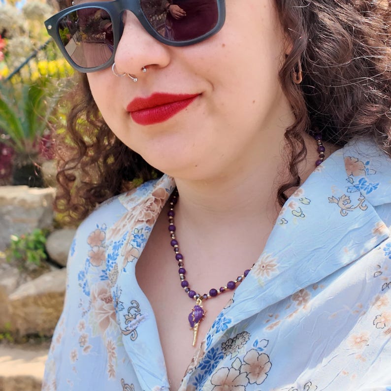 The necklace worn against a pale blue floral shirt by water, the row of small purple beads sitting close to the neck with the gold-tipped purple pendant below.