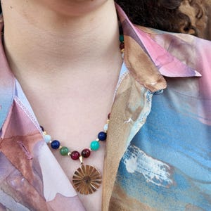 The necklace worn open-collar in daylight, the multicoloured bead strand crossing the collarbones and the gold sunburst disc resting in the neckline between two deep wine-red beads.