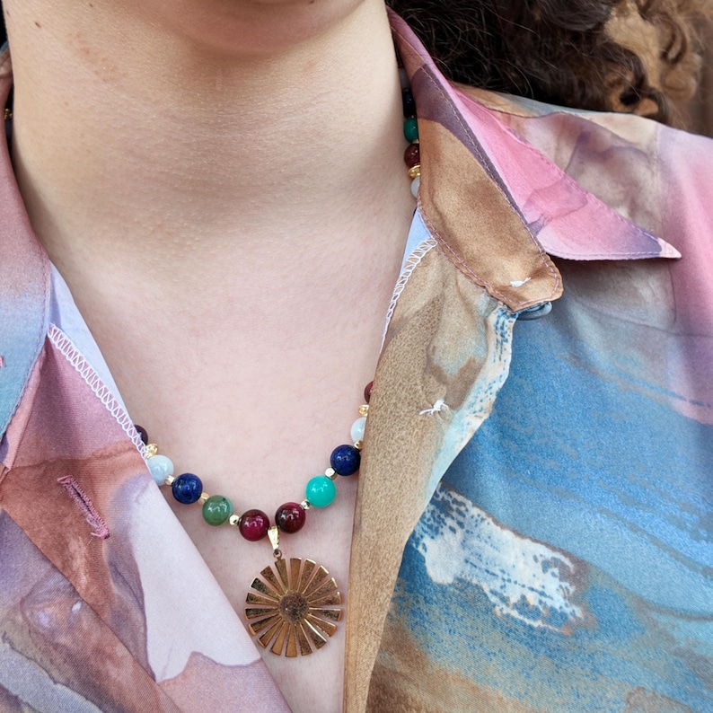 The necklace worn open-collar in daylight, the multicoloured bead strand crossing the collarbones and the gold sunburst disc resting in the neckline between two deep wine-red beads.