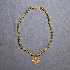 Flat-lay on grey-blue cloth of a short green beaded necklace, small round stone beads alternating with slim gold-tone tubes, ending in a daisy pendant with a round green centre; lobster clasp and extender chain at the top.