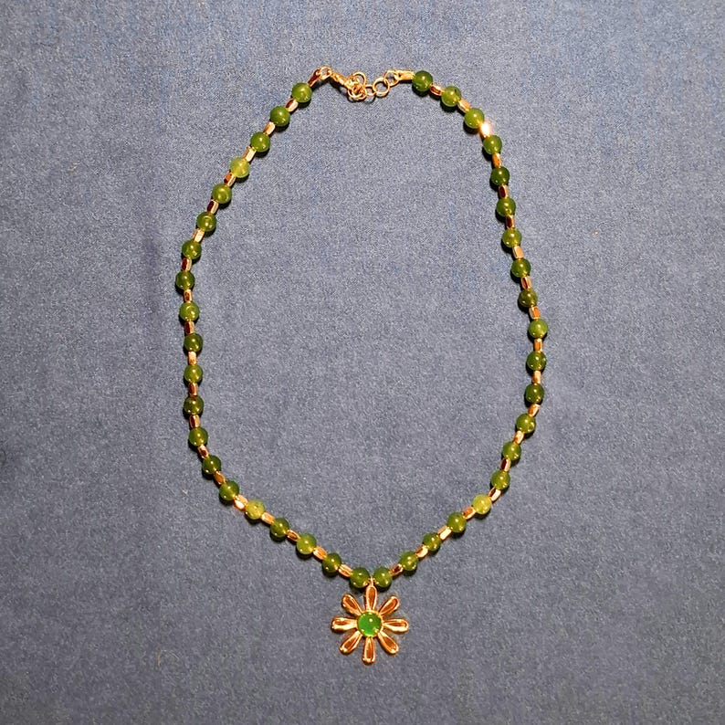Flat-lay on grey-blue cloth of a short green beaded necklace, small round stone beads alternating with slim gold-tone tubes, ending in a daisy pendant with a round green centre; lobster clasp and extender chain at the top.