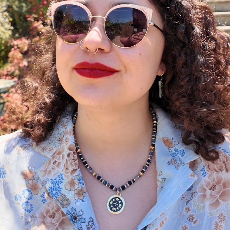 Daylight close view of the strand over a pale floral shirt collar, deep blue beads broken up by gold rondelles and clear rhinestone rings, the medallion resting in the open V of the shirt.