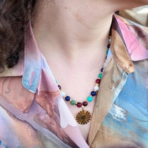 Close view of the strand on the neck, showing the repeating run of navy, milky cream, green and wine-red round beads with tiny gold-tone cubes between them and the ridged gold disc below.