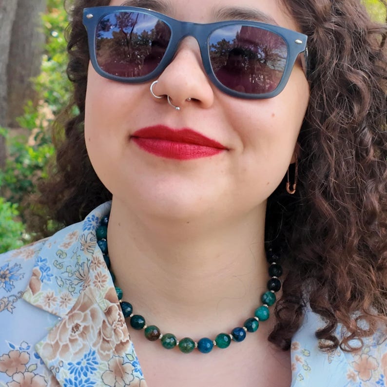 The beaded strand worn snug at the base of the neck over a pale blue floral blouse, its green and teal faceted beads catching afternoon daylight with small bright spacers between them.