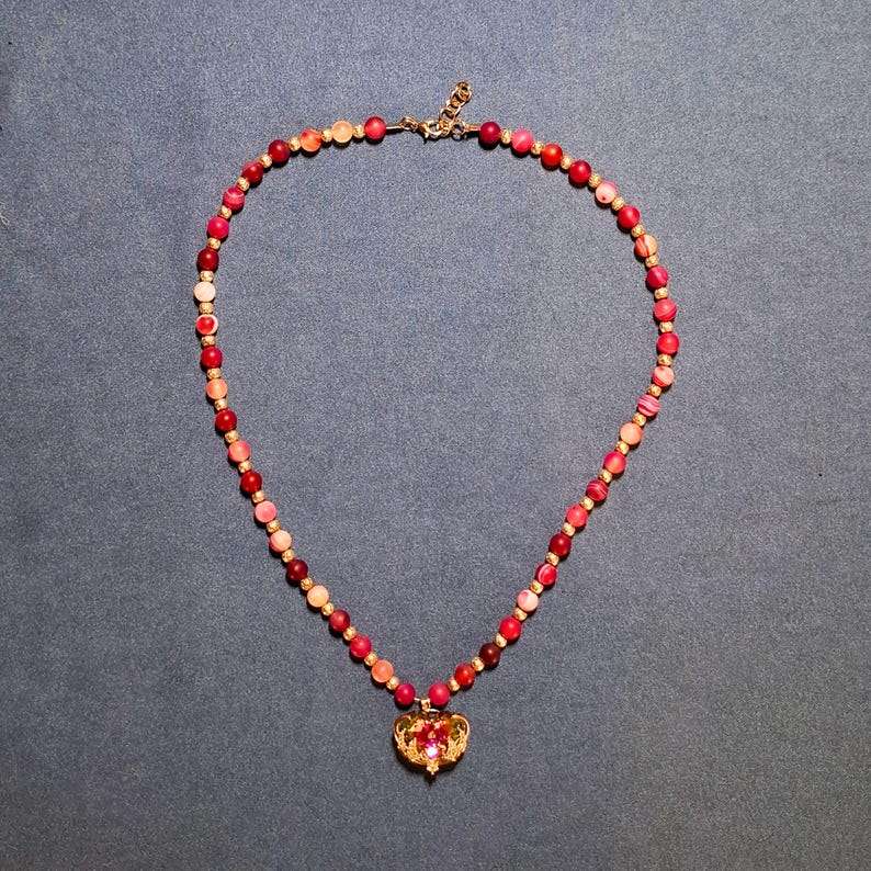 The necklace laid in a full circle on grey-blue cloth: matte round beads shading from pale peach through bright pink to deep red, a small ornate spacer between each one, and a heart pendant with a faceted iridescent stone at the front.