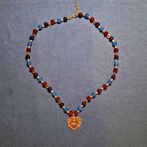 Beaded necklace laid in an oval on grey-blue cloth: polished 8 mm rounds in cherry red, deep plum and bright blue with gold-tone spacers, a gold heart pendant with an eye motif at the bottom centre.