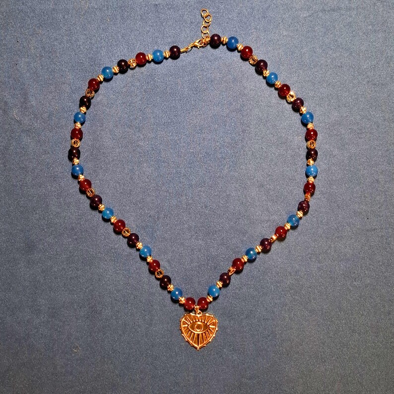 Beaded necklace laid in an oval on grey-blue cloth: polished 8 mm rounds in cherry red, deep plum and bright blue with gold-tone spacers, a gold heart pendant with an eye motif at the bottom centre.