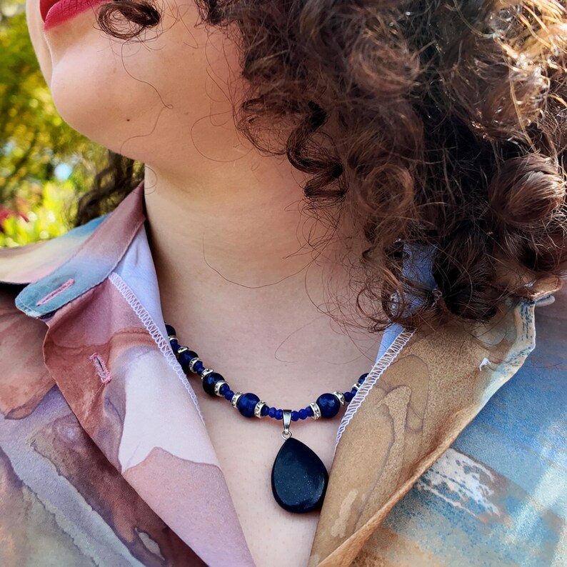 Close view of the navy bead strand worn at the base of the throat, the round beads and sparkling spacers following the collarbone, with the dark teardrop pendant resting in the open neckline of a pastel printed shirt.