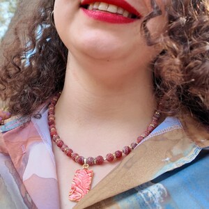 Angled outdoor view with curls falling over the strand: the frosted red beads and gold spacers curve under the collar and the gold-edged coral pendant sits centred at the opening of the shirt.