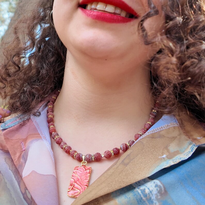 Angled outdoor view with curls falling over the strand: the frosted red beads and gold spacers curve under the collar and the gold-edged coral pendant sits centred at the opening of the shirt.