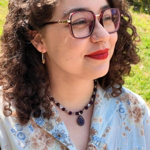Worn outdoors in daylight, the deep purple strand rests along the collarbone with the teardrop pendant hanging just below the hollow of the throat; small pale spacers catch the sun between the round beads.