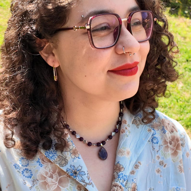 Worn outdoors in daylight, the deep purple strand rests along the collarbone with the teardrop pendant hanging just below the hollow of the throat; small pale spacers catch the sun between the round beads.