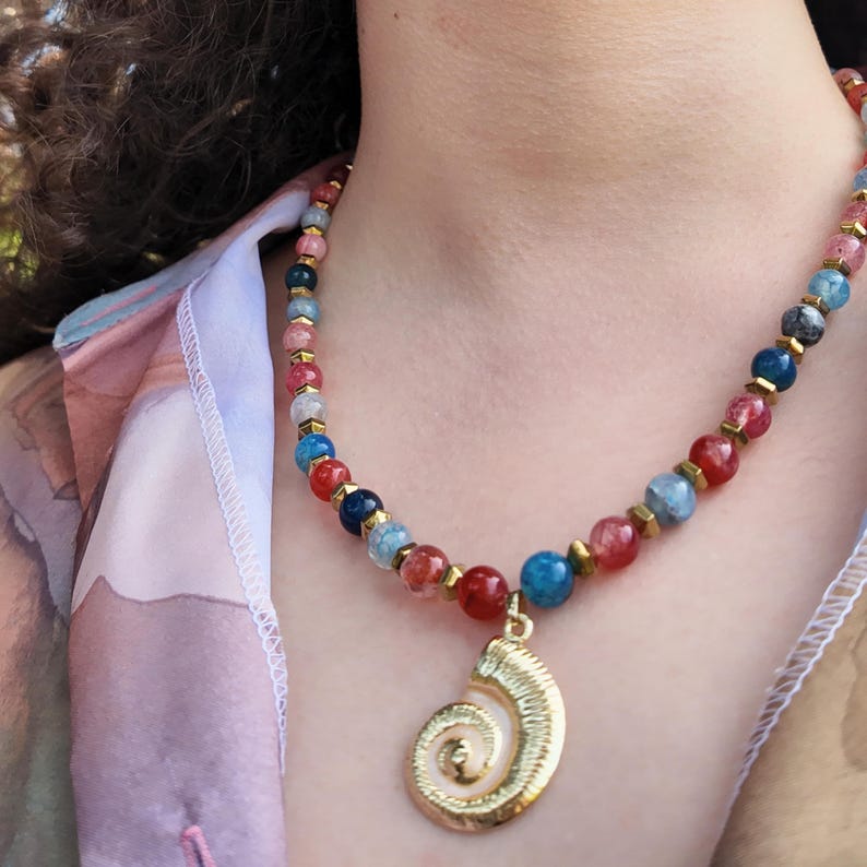 The beaded strand sits snug around the base of the neck over an open pastel shirt, red, blue and pale grey beads catching daylight, gold spiral shell pendant hanging at the centre of the chest.