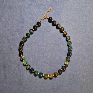 A single strand of faceted 8 mm stone beads laid in an oval on grey-blue cloth, running from near-black through deep teal and forest green to honey amber, with a tiny metal spacer set between every bead.