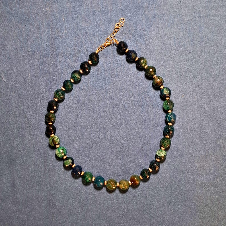 A single strand of faceted 8 mm stone beads laid in an oval on grey-blue cloth, running from near-black through deep teal and forest green to honey amber, with a tiny metal spacer set between every bead.