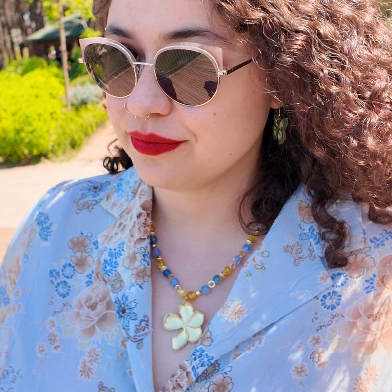 Side view outdoors: the beaded row curves under the hair and around the neck, blue and yellow beads shown at wearing scale, with the flower pendant sitting centred at the chest.