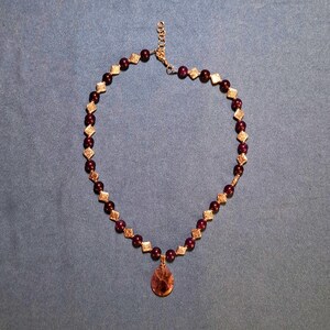 Flat-lay of a beaded necklace on blue-grey fabric: round dark plum stone beads alternate with small diamond-shaped patterned metal spacers, a smooth teardrop pendant hangs at the front centre, and a short extender chain with a lobster clasp closes