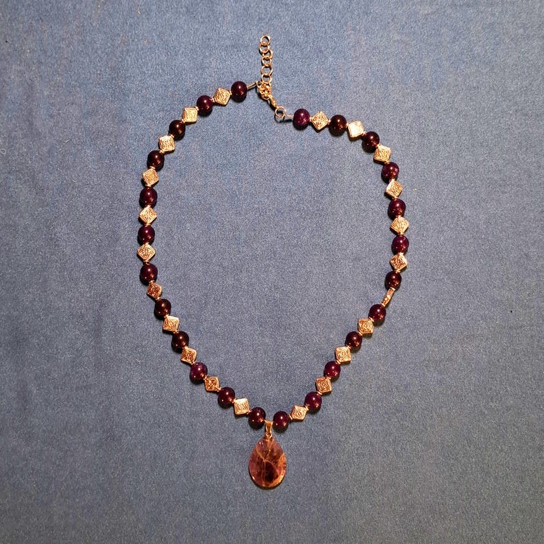 Flat-lay of a beaded necklace on blue-grey fabric: round dark plum stone beads alternate with small diamond-shaped patterned metal spacers, a smooth teardrop pendant hangs at the front centre, and a short extender chain with a lobster clasp closes