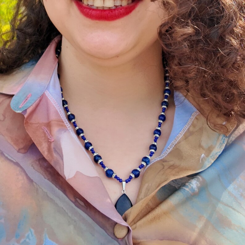 Side angle in sunlight showing the necklace running down the neck and over the shoulder of a soft pink and tan shirt, round royal blue beads catching the light, the teardrop pendant hanging against the chest.