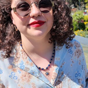 Close view of the necklace on the collarbone, showing small round royal-blue beads separated by bright rondelles and a few dark discs, with the gold sun pendant and its blue centre stone below.