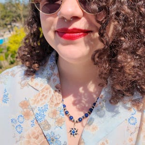 Three-quarter daylight view of the blue bead strand curving along the collarbone, the tiny rondelles flashing white in full sun, the sun pendant hanging just inside the open collar of a pale floral shirt.