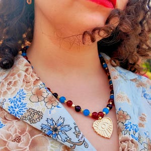 The necklace worn over a pale floral shirt in daylight, the red, plum and blue beads sitting around the base of the neck and the gold heart pendant with its eye motif resting just below the collarbones.