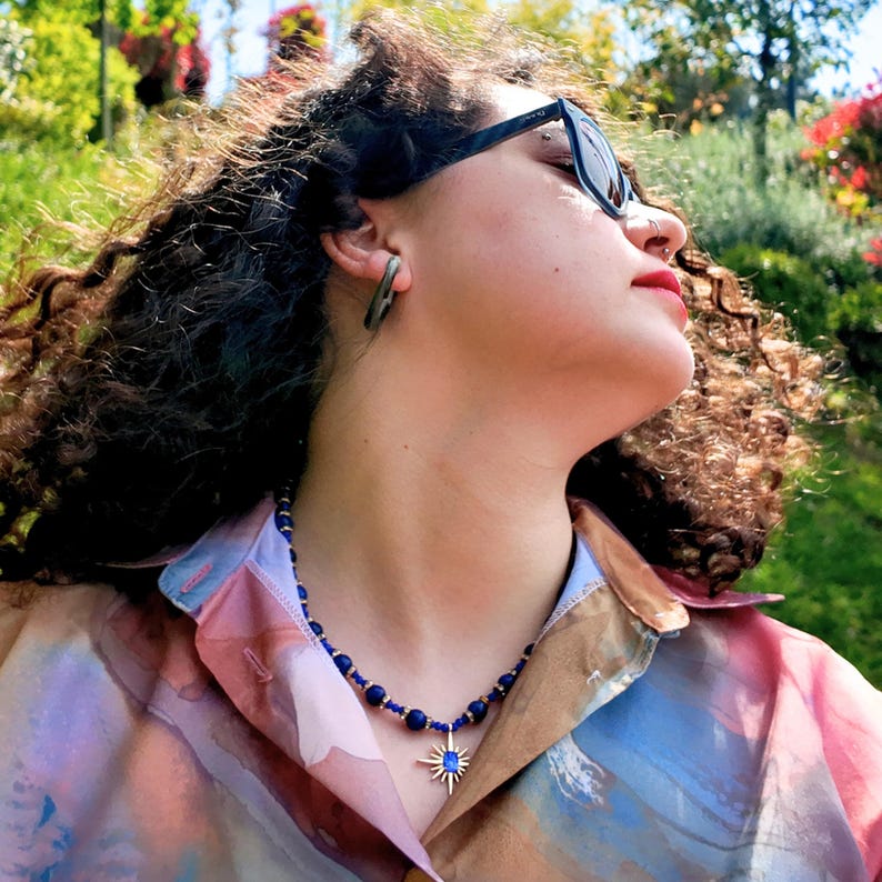 Close view over an open pastel shirt: navy round beads and small gold spacers curve under curly hair, with the gold starburst and its blue centre stone at the front.