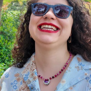 Side-on view across the collarbone showing deep pink and plum beads separated by small metallic rounds, the heart pendant and its rainbow-flashing stone sitting at the top of the chest.